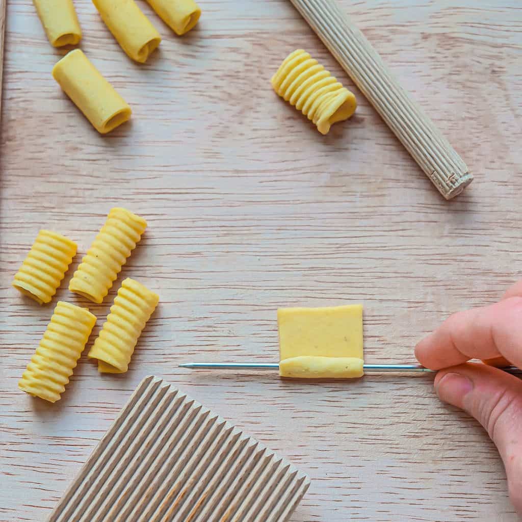 An overhead view of a small square of pasta being rolled onto a metal skewer to form a tube shape.