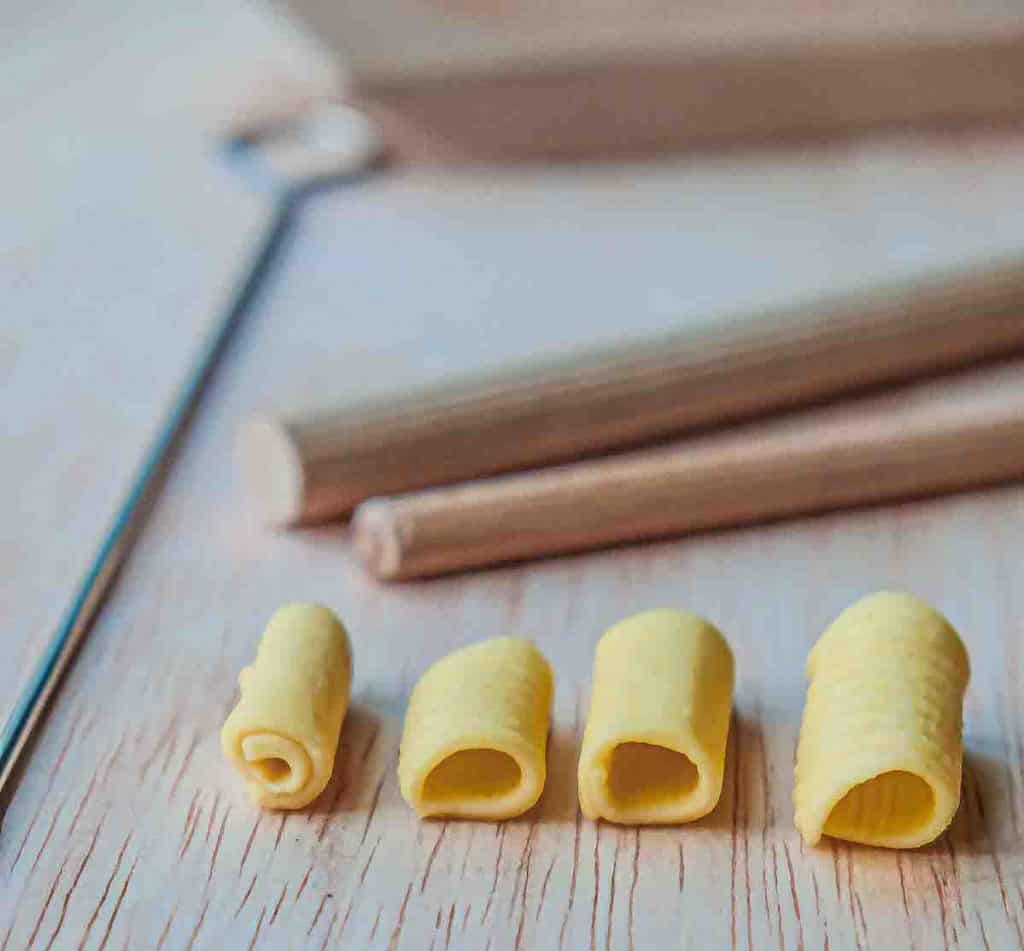 Different widths of tube shaped pasta with different sized wooden dowels and a skewer in the background.