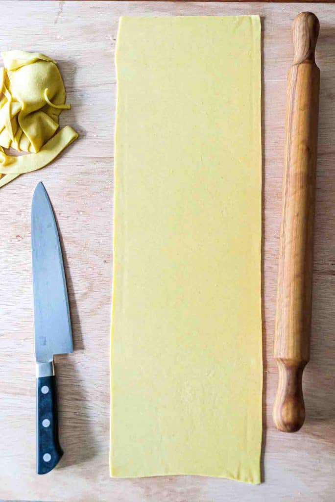 A smooth sheet of semolina pasta dough that's been rolled out with a rolling pin sitting to its right and then trimmed into a rectangle with a chef's knife sitting to its left.