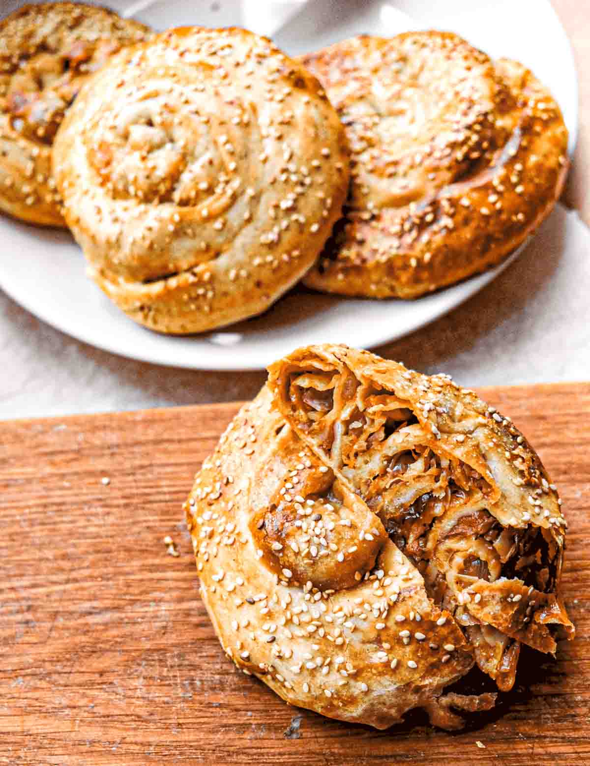 In the foreground there is a cabbage phyllo spiral that has been cut in half to show the layers of dough and cabbage on the inside. In the background there is a bowl of golden phyllo spiral buns with sesame seeds on top.