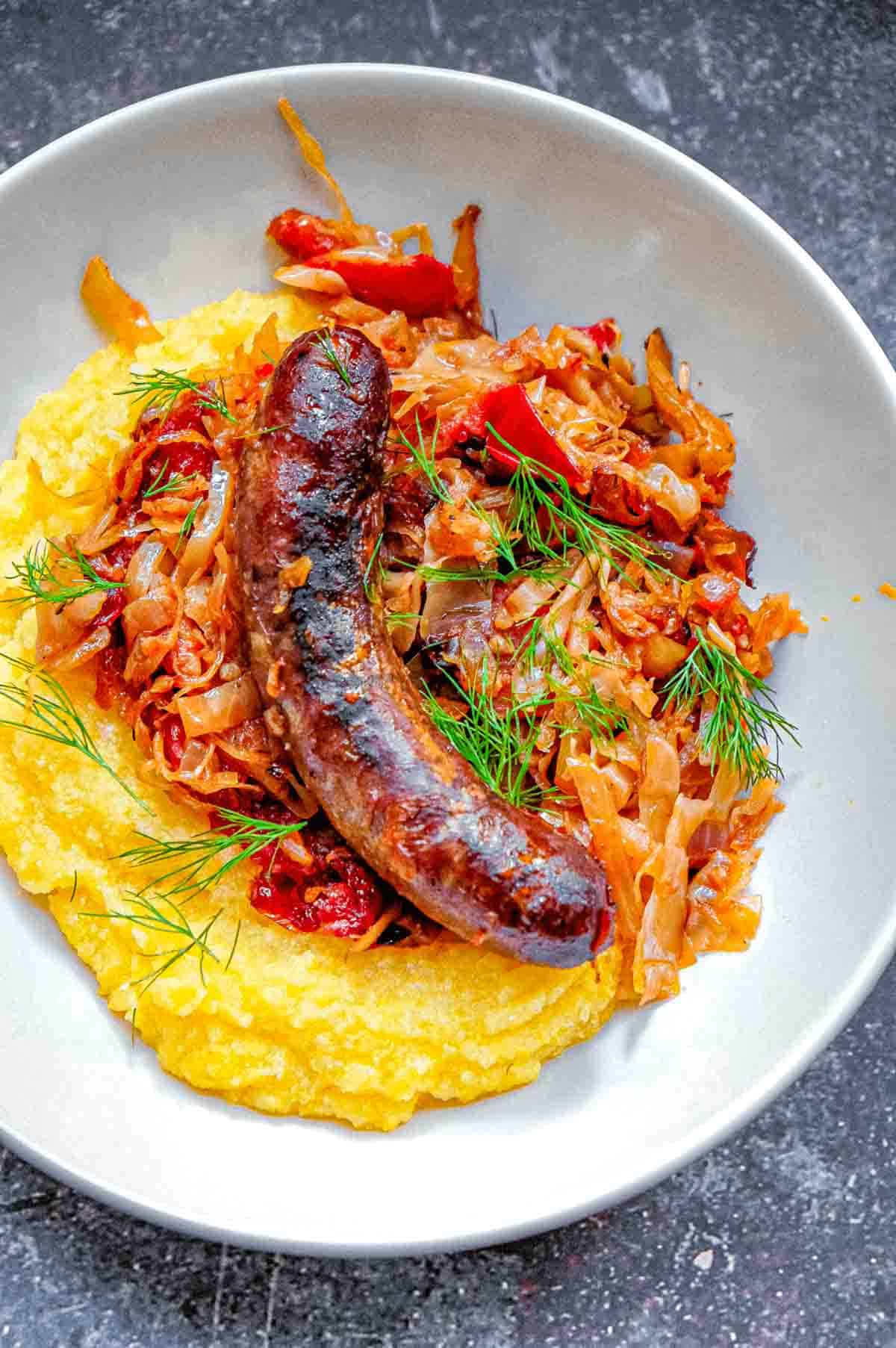 A bowl of slow roasted Romanian sauerkraut with smoked pork sausage over creamy yellow polenta and with a few sprigs of dill as garnish.