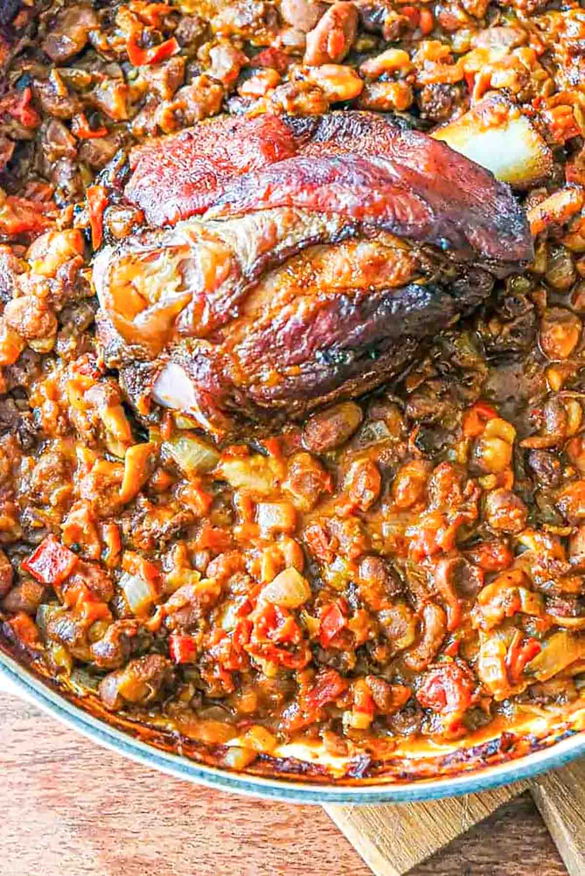 A whole smoked ham hock with crispy roasted skin laying on top of a big pot of baked beans cooked with tomato.