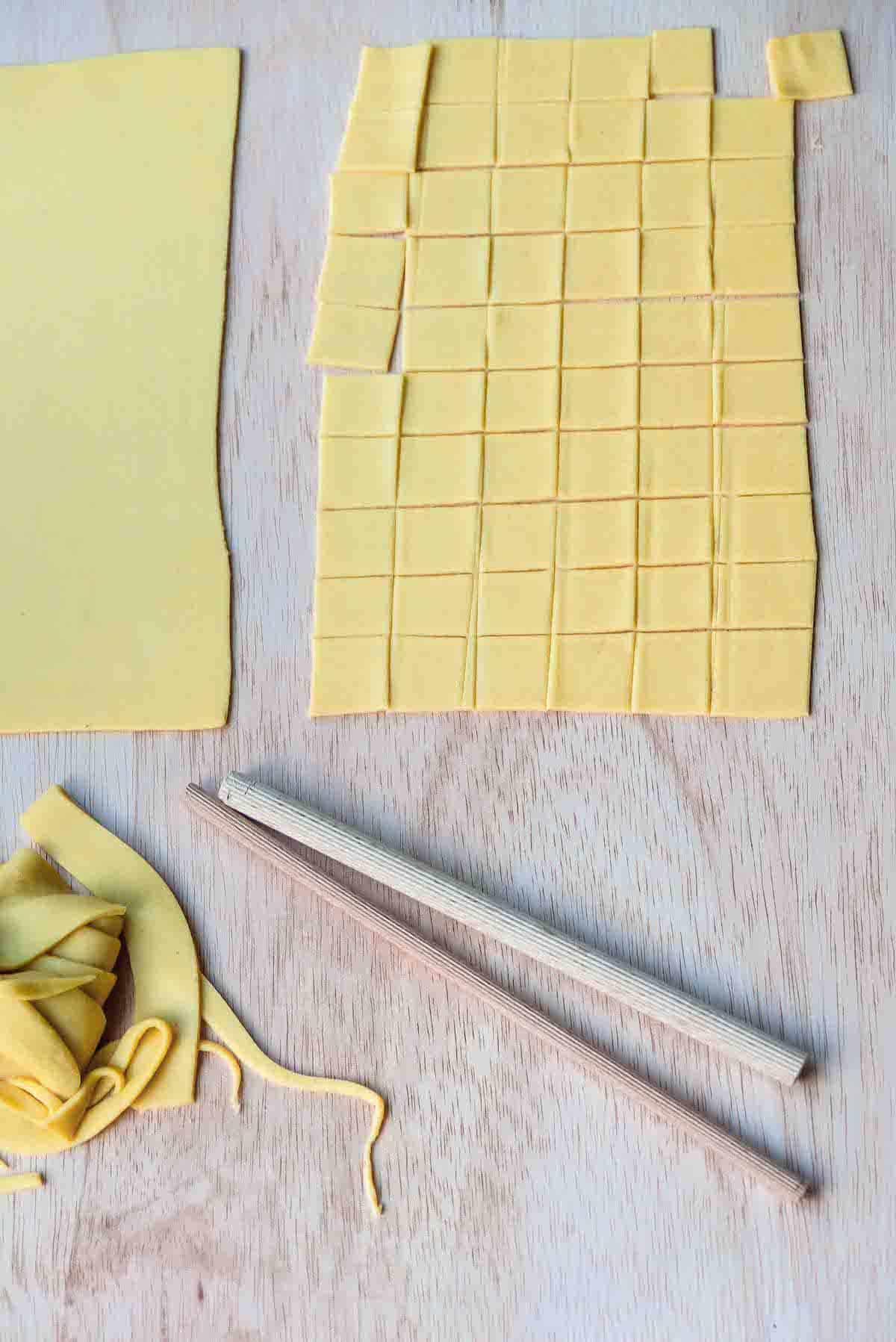 A sheet of pasta dough cut into even squares and below that are two wooden dowels of different sizes.