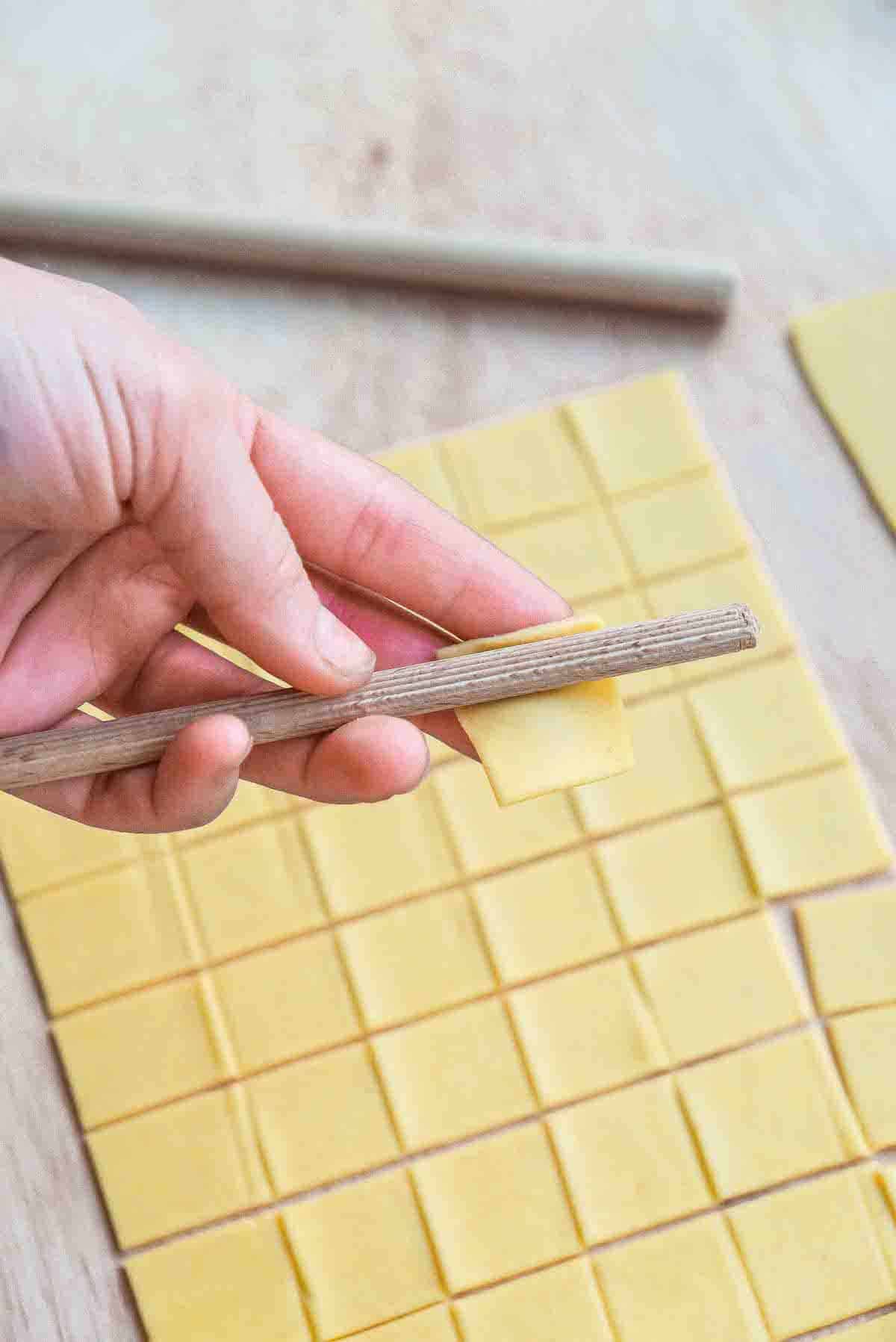 Rolling a square piece of pasta dough onto a ridged wooden dowel to make a macaroni shape.