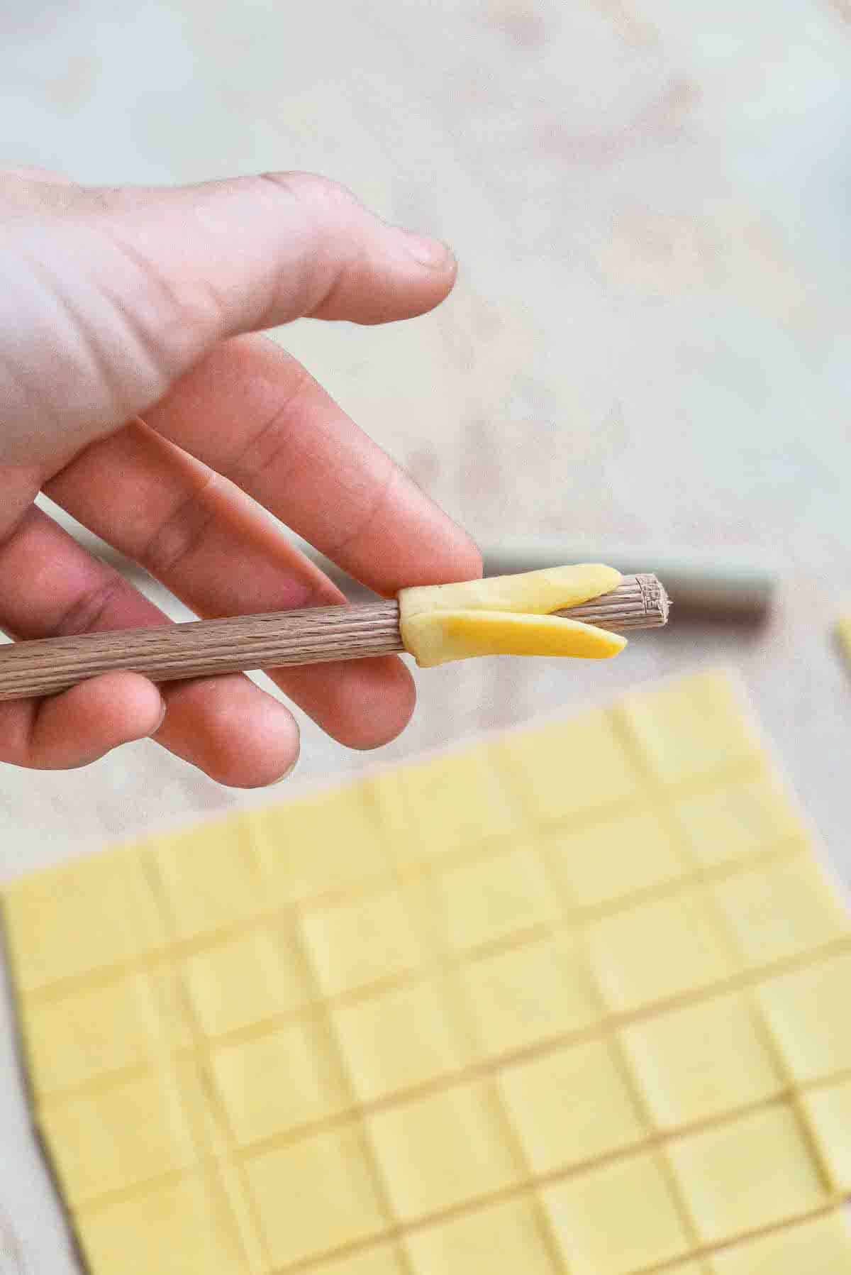A square of pasta dough almost fully rolled into a tube over a ridged wooden dowel.