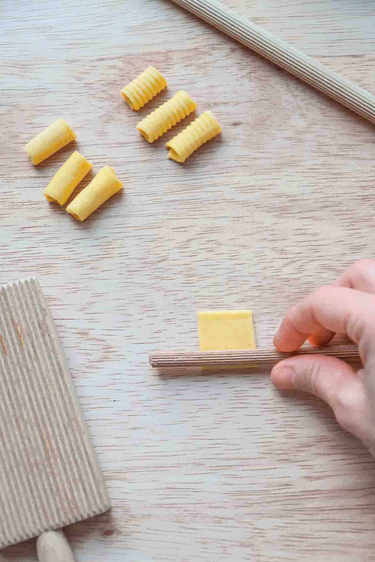 Pressing a ridged wooden dowel into a square of pasta dough in order to make a macaroni shape like the 6 macaronis lying just above the square of dough and dowel.