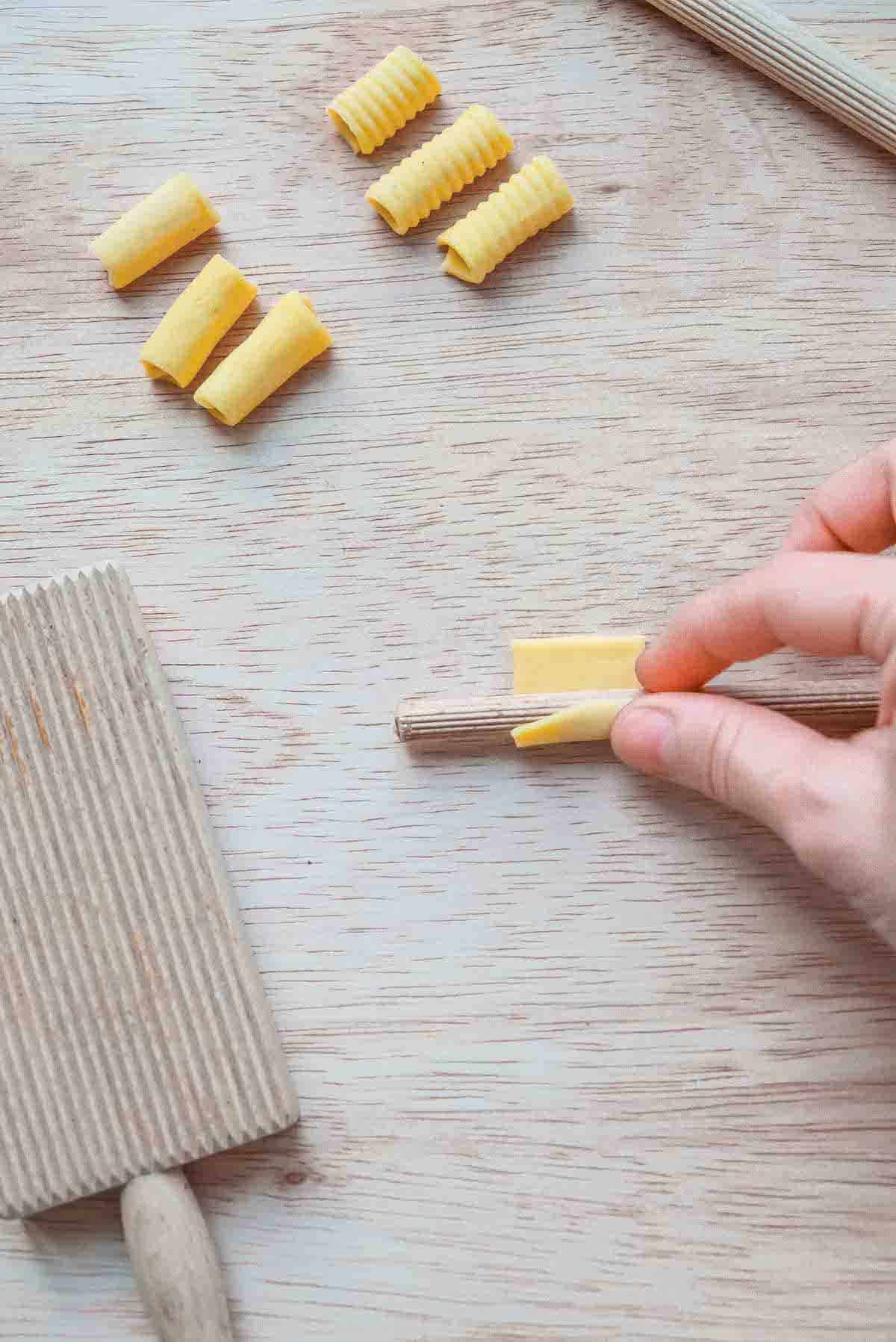 A square of pasta dough is being gently pressed onto a ridged wooden dowel in order to roll it into a tube.