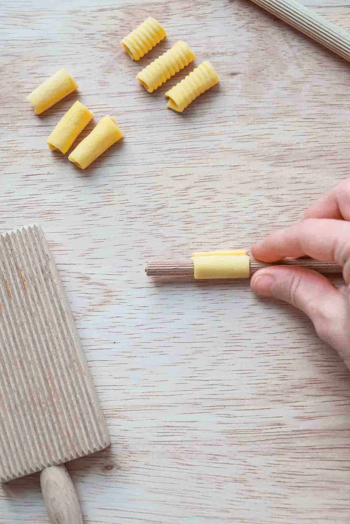 A square of pasta dough has been rolled onto a ridged wooden dowel into a tube shape.