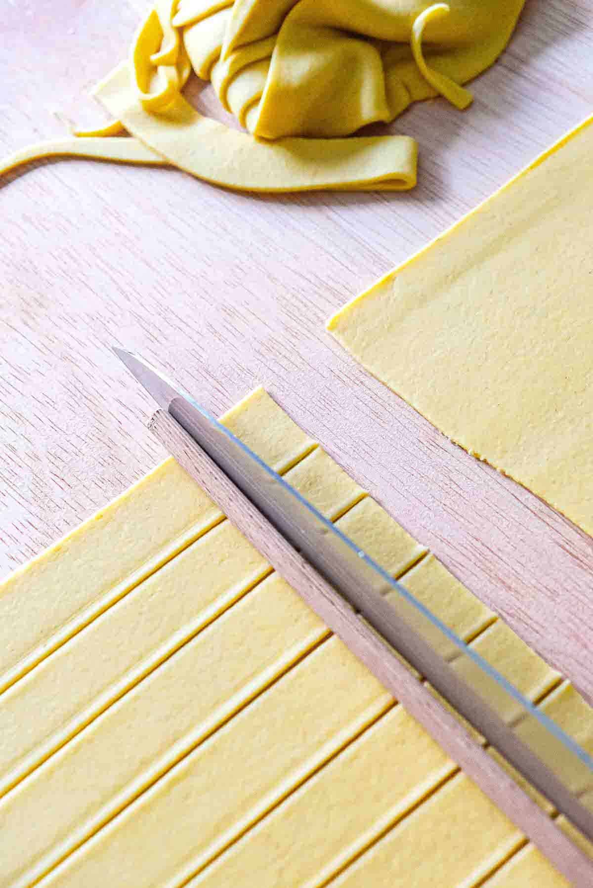 Cutting a sheet of pasta dough into squares using a large chef's knife and a wooden dowel as a guide.