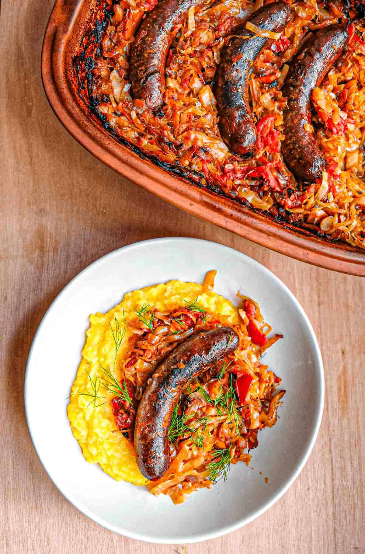 An overhead view of a bowl of slow cooked polenta with sausage on top of polenta. Above the plate there is a clay pot full of cooked cabbage and roasted sausages.