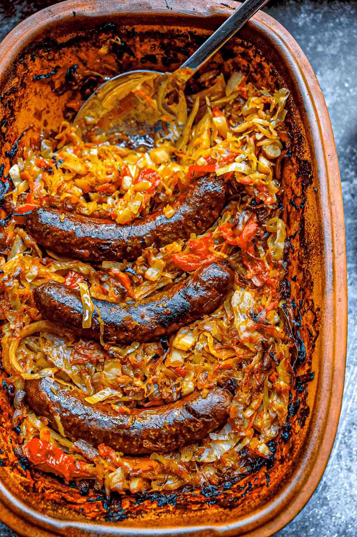 An overhead view of a large clay pot full of slow roasted cabbage and sausages.