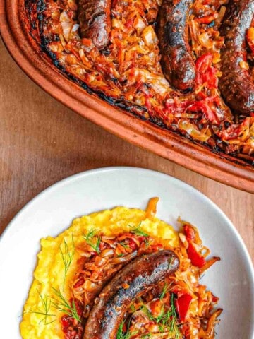 An overhead view of a plate of slow roasted sauerkraut over polenta with sausage and a dill garnish. Above it you can see the clay pot full of cabbage and sausages.