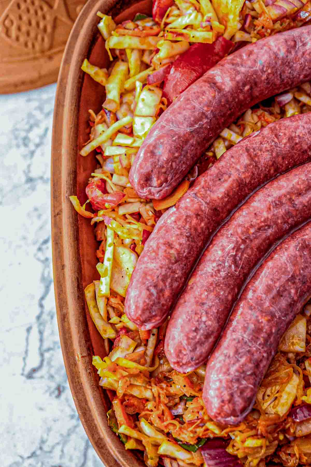 Uncooked smoked pork sausages on top of uncooked cabbage and vegetables in a clay pot.