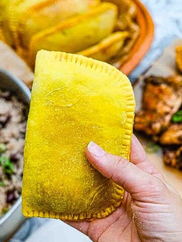 A Jamaican beef patty being held up in my hand against a backdrop of other Jamaican dishes like rice and peas and jerk chicken.