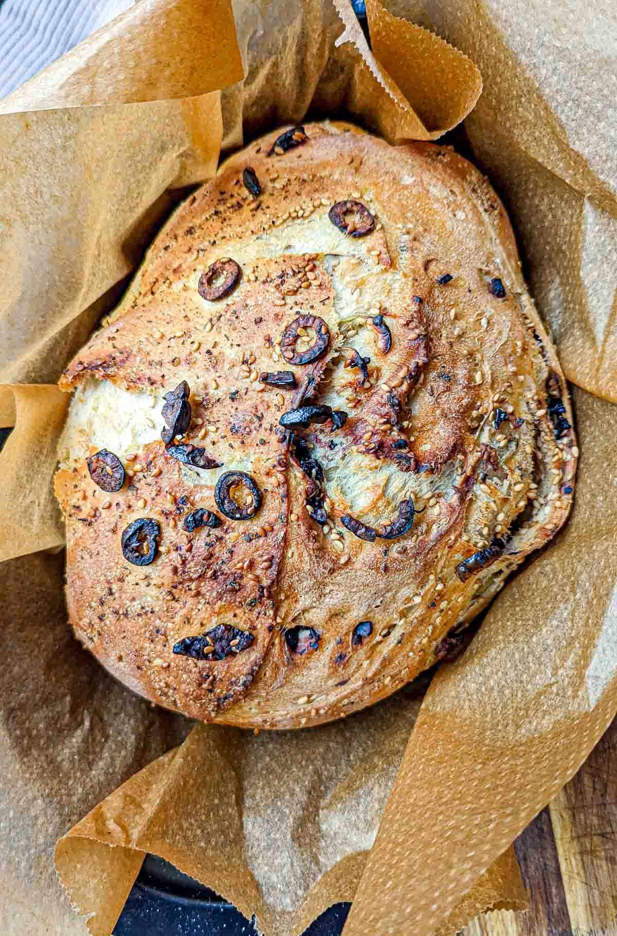 A golden-brown sesame and olive bread loaf on parchment paper.