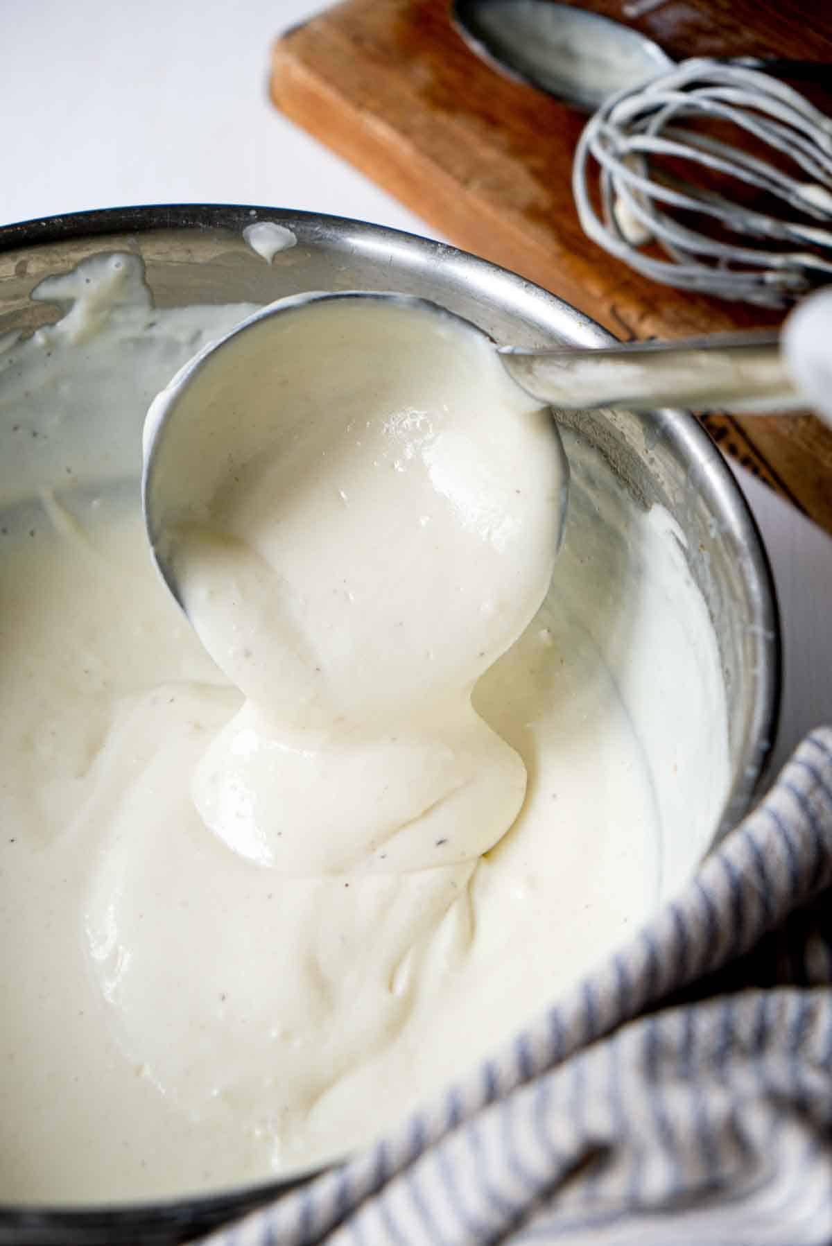 Thick bechamel sauce pouring from a ladle into a stainless steel sauce pan.