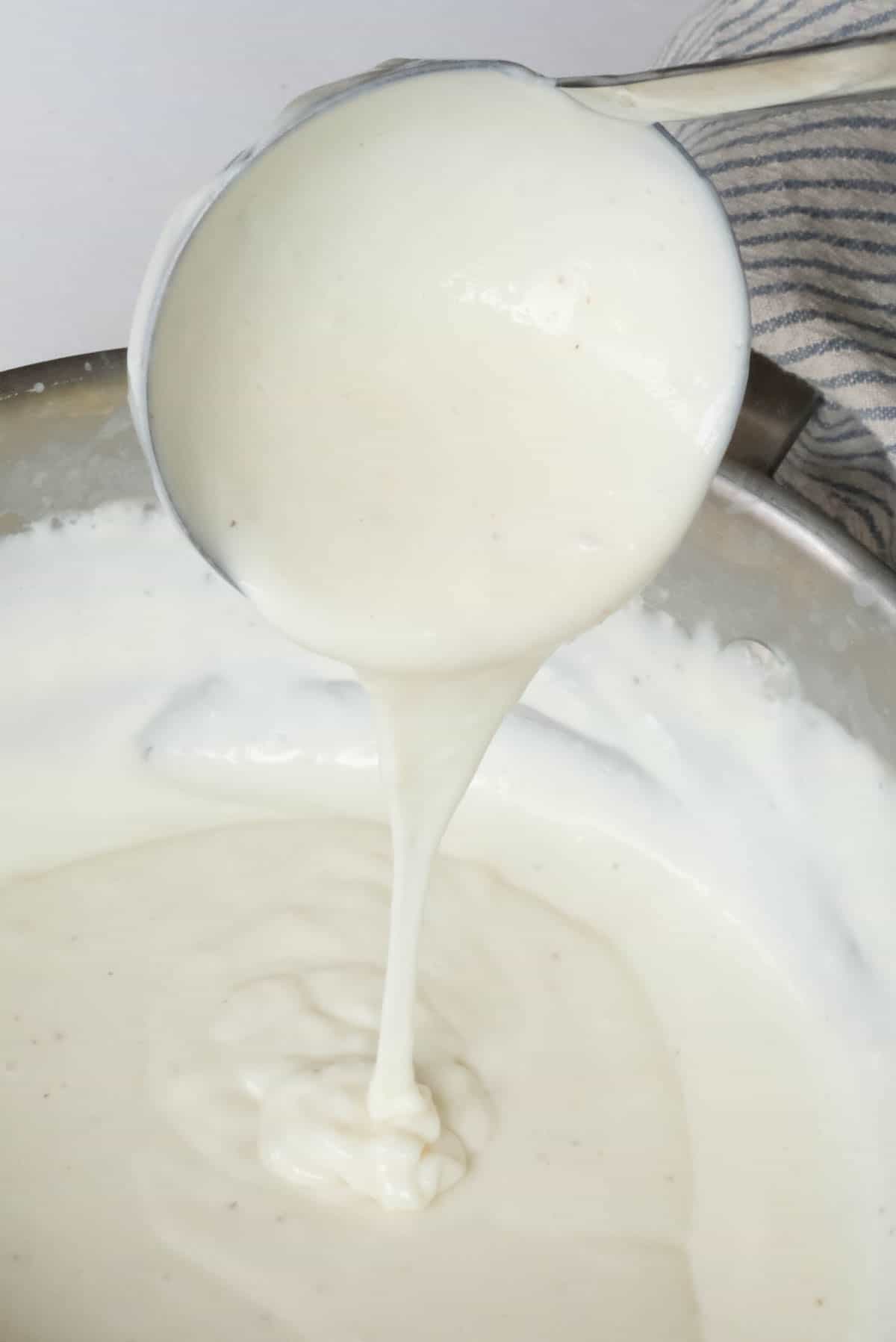 Bechamel immediately after preparing showing a thin stream of sauce pouring from a ladle into a pot.