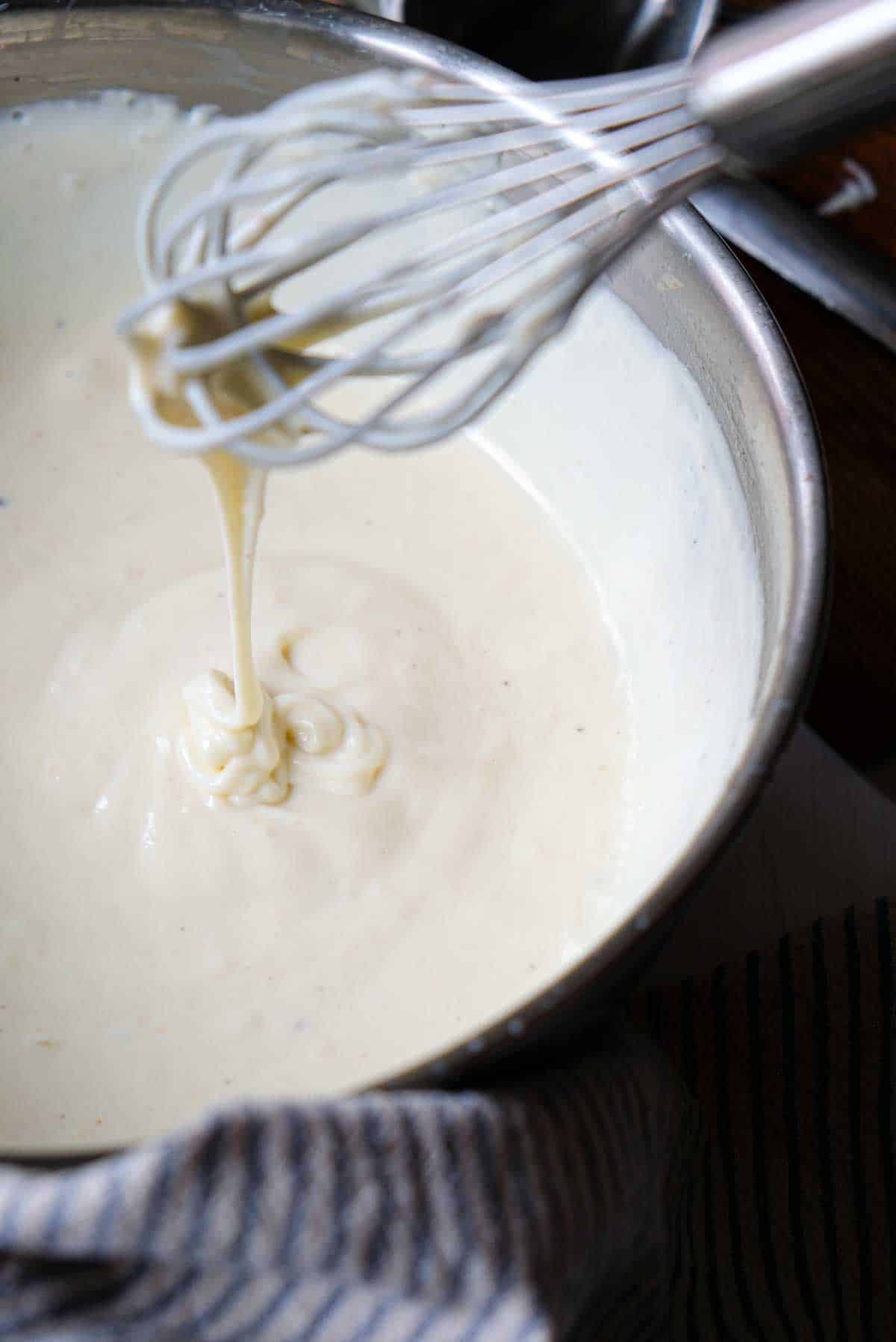 Bechamel sauce dripping from a wire whisk into a stainless steel pot.