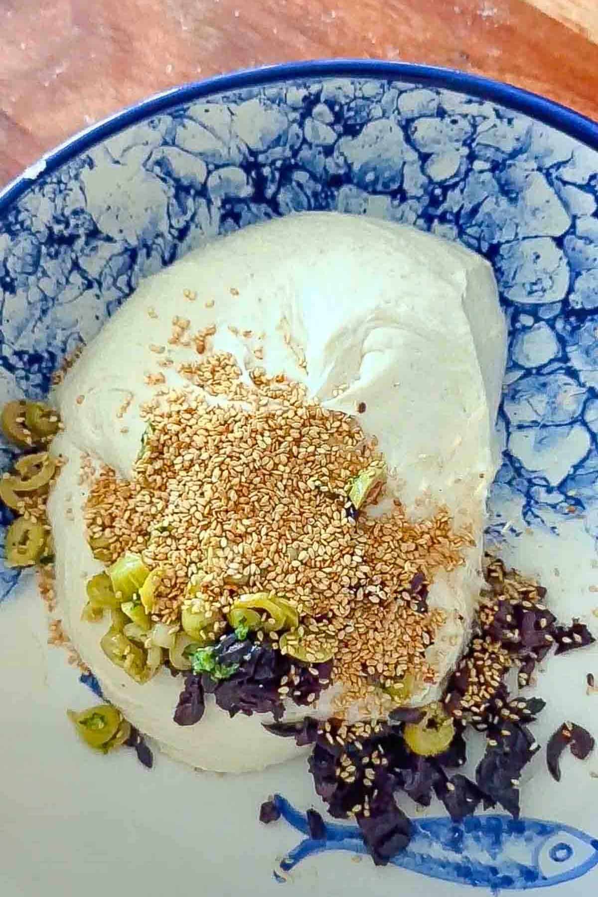 A large bowl containing bread dough, sesame seeds, sliced green olives and sliced black olives.