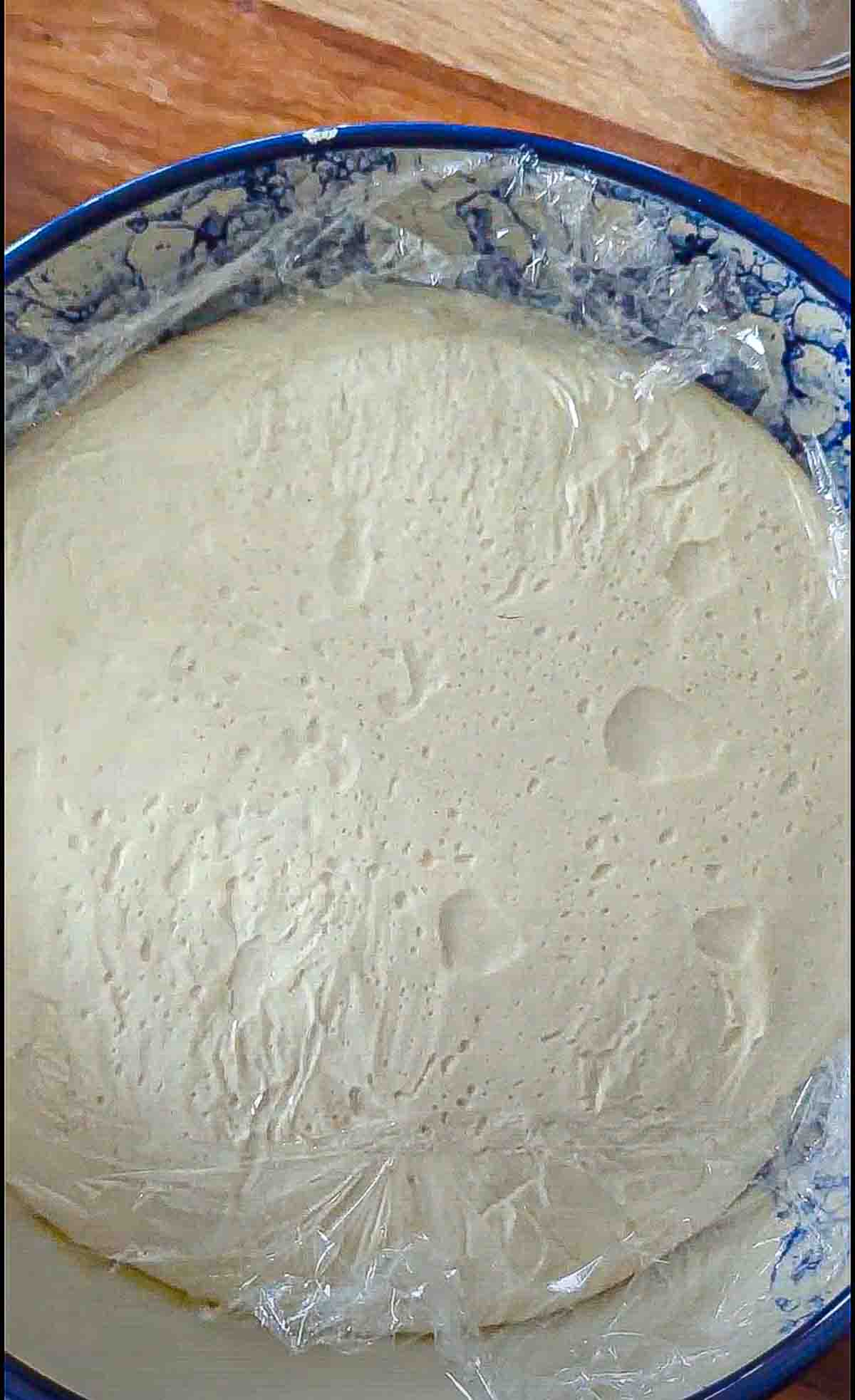 A large bowl containing risen bread dough that's expanded all the way to the edges of the bowl, is covered in plastic wrap and has some bubbles on its surface.