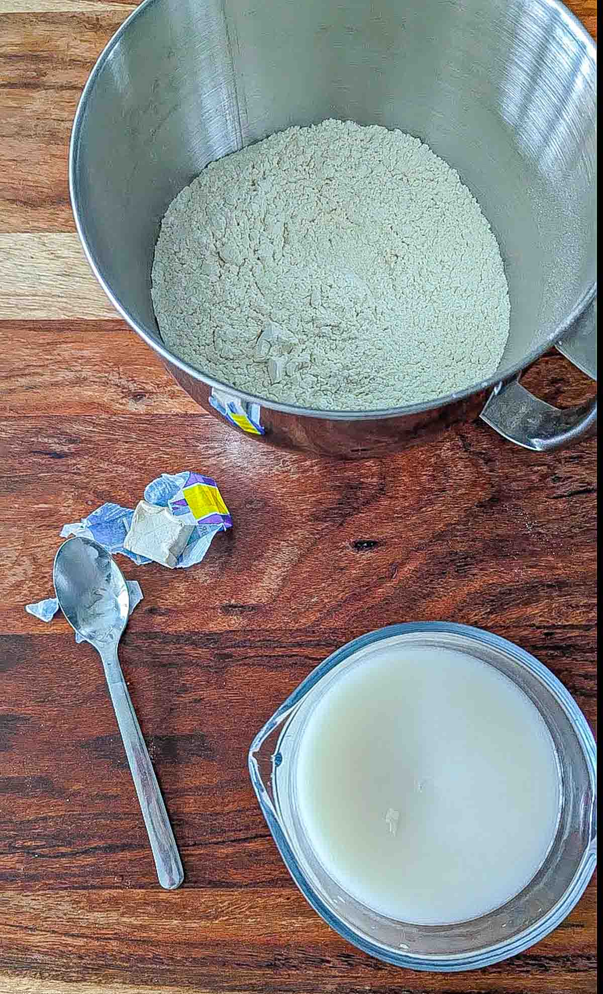 The bowl of a Kitchenaid stand mixer with flour, a small package of fresh yeast, and a glass measuring cup with water and fresh yeast blooming.