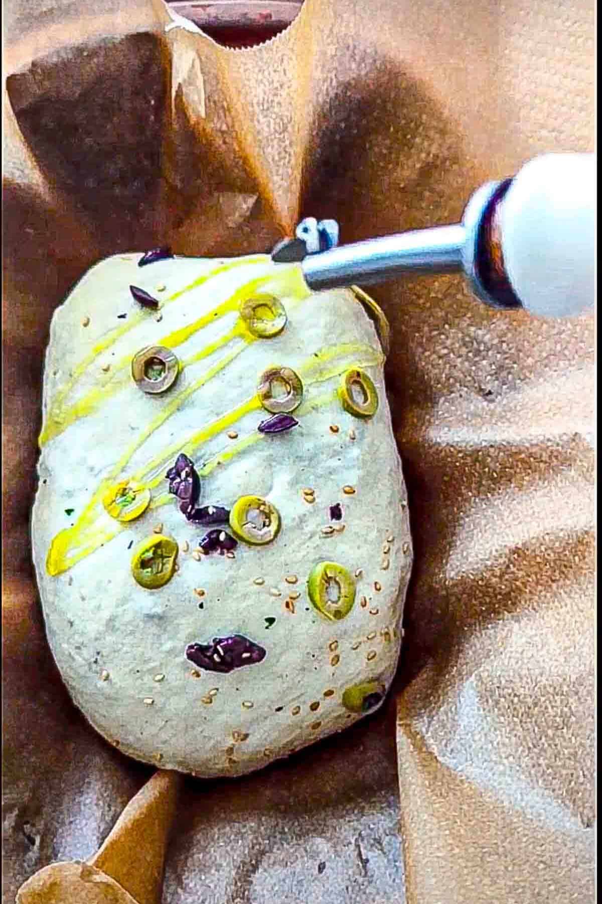 A parchment paper lined loaf pan containing bread dough shaped into an oval boule topped with green olives, black olives and sesame seeds and being drizzled with olive oil.