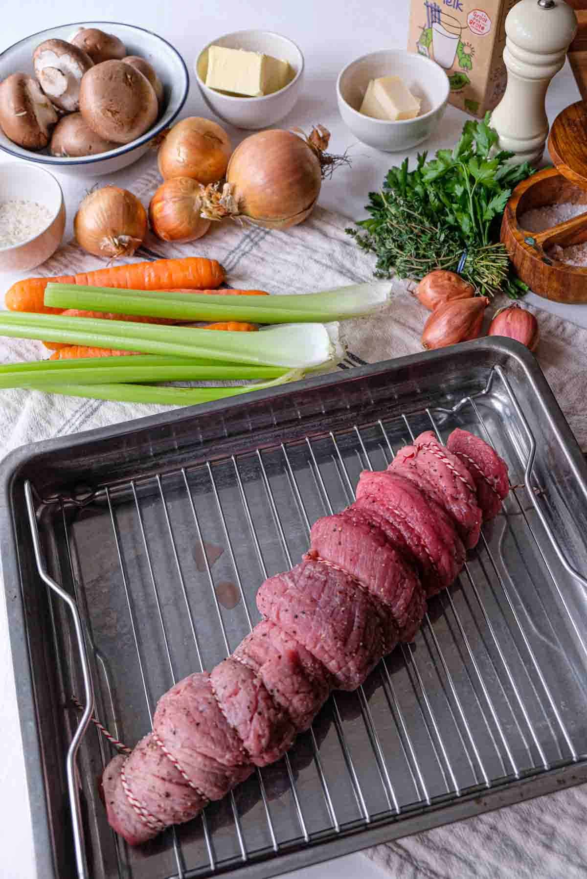 In the foreground a trussed veal loin in a roasting pan and in the back the rest of the ingredients for Veal Prince Orloff including onions, mushrooms, roasting vegetables, millk, and butter.