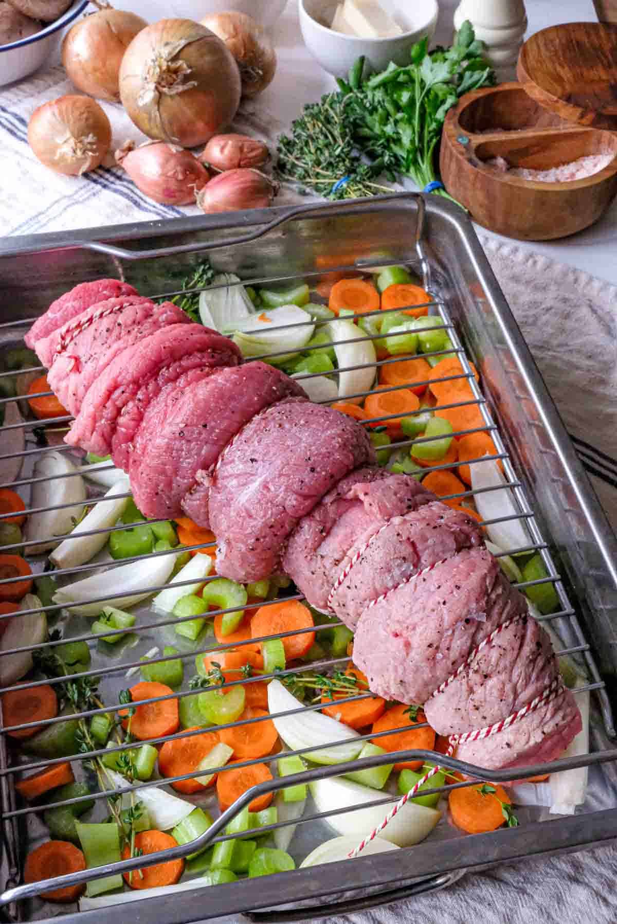 A trussed veal laying on top of a rack in a roasting pan with carrots, celery, onions and fresh thyme.