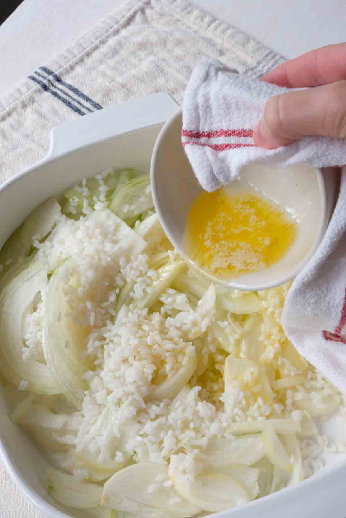 A casserole dish with onion and rice in it being drizzled with melted butter.