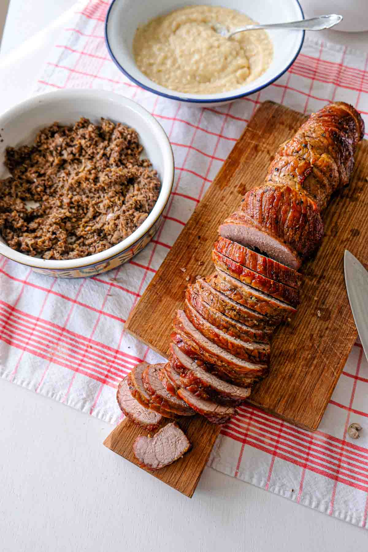 Roasted veal being cut into thin slices with mushrooms duxelles and onion soubise in separate dishes nearby.