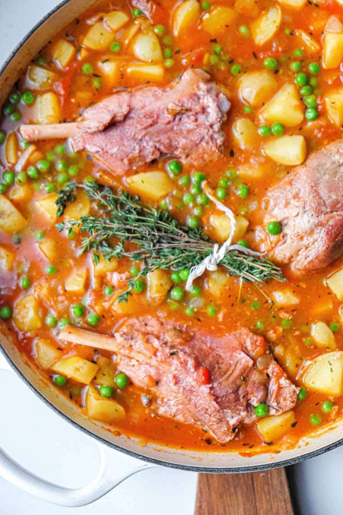 Close up view of the ingredients in potato stew before cooking. You can see potatoes, peas, a bundle of fresh thyme, and 3 large ham hocks in a base of crushed tomatoes and seasonings.