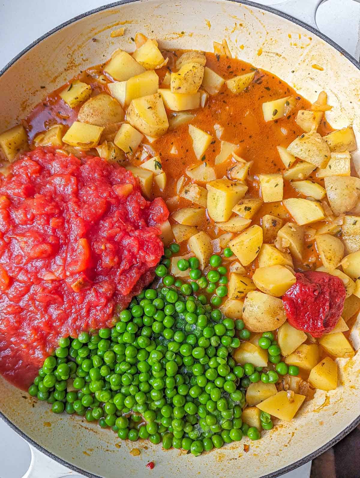 A large enameled cast iron pot containing lightly cooked cubed potatoes, crushed tomatoes, tomato paste and green peas.