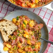 A plate of cooked potato stew with peas and smoked ham along with a slice of bread.