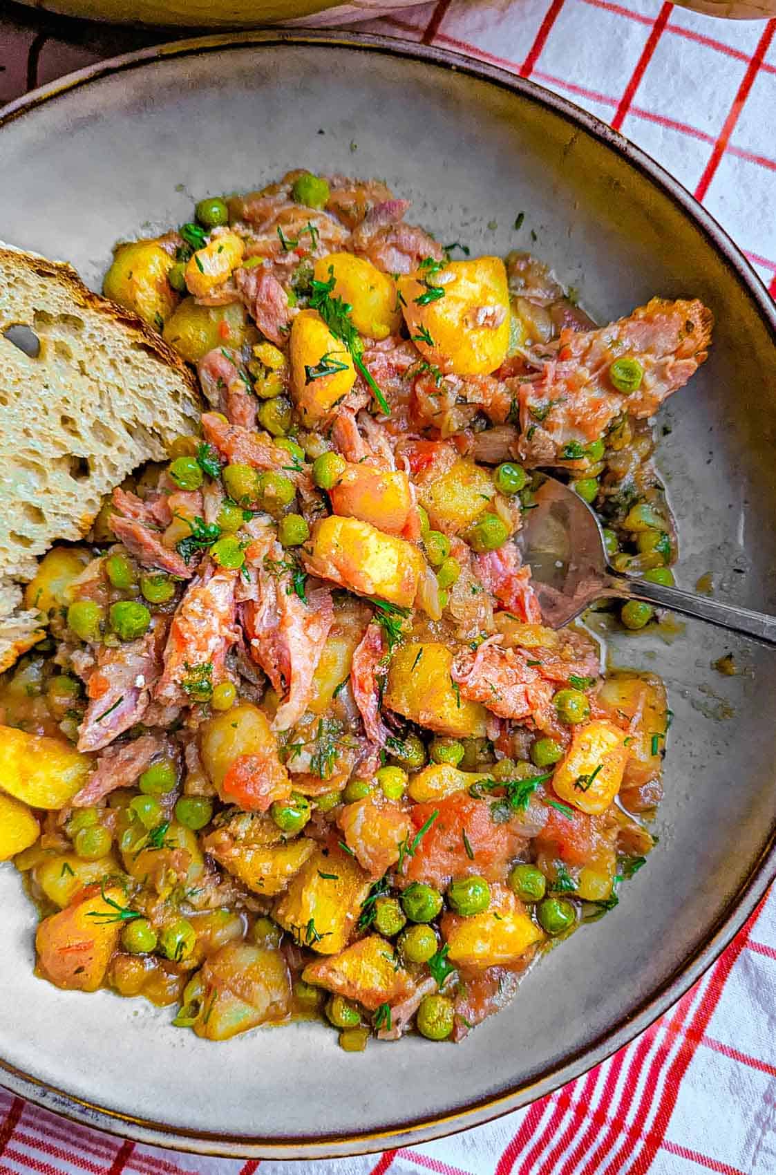 A plate of cooked potato stew with peas and smoked ham along with a slice of bread.