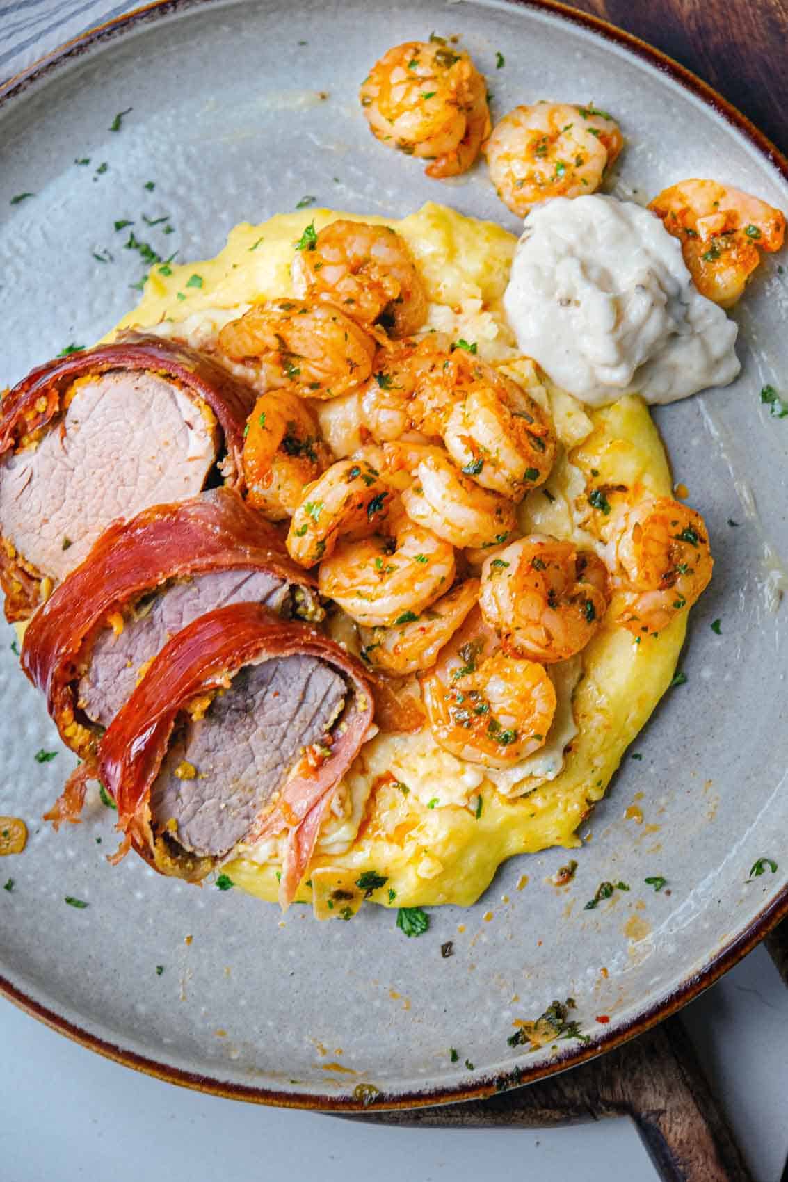 a plate of creamy polenta with prosciutto wrapped pork tenderloin, red pepper shrimp and roasted garlic parmesan aioli