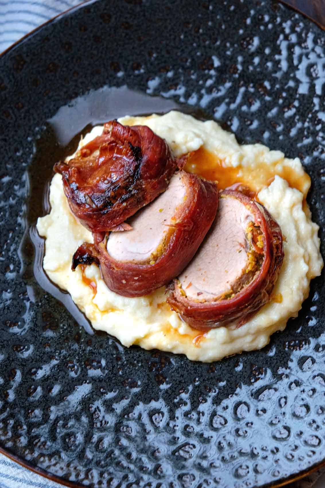 prosciutto wrapped pork tenderloin sliced on a plate overtop creamy polenta
