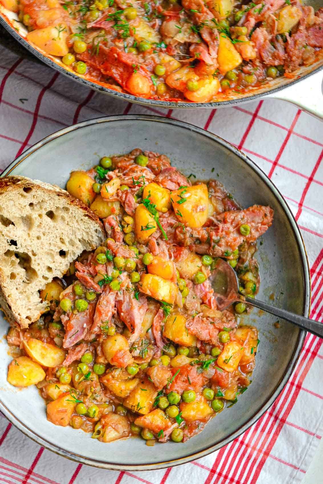 A plate of cooked potato stew with peas and smoked ham along with a slice of bread.