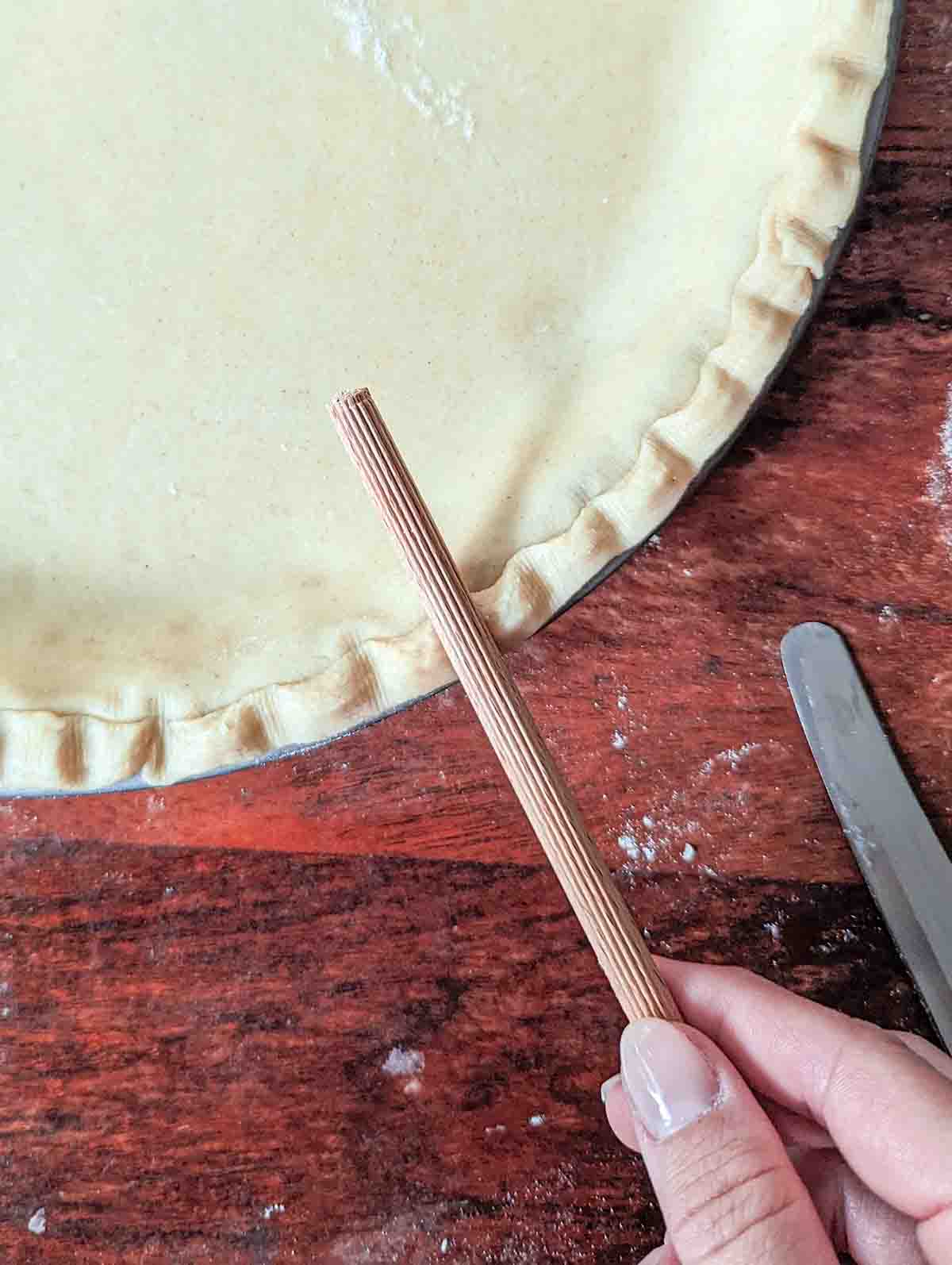 A close up look at the edge of the pie crust which has been lined with strips of pie dough that are being lightly pressed with a ridged wooden dowel to create a crimped edge on the pie.