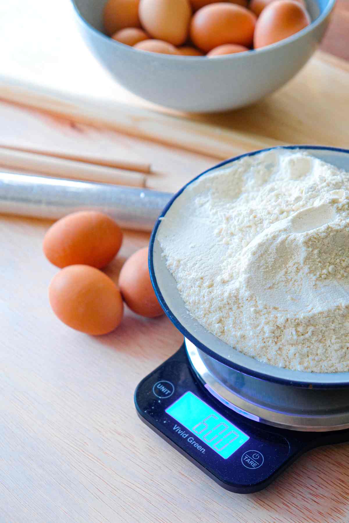 Flour in a bowl on a kitchen scale with some eggs and rolling pins in the background.