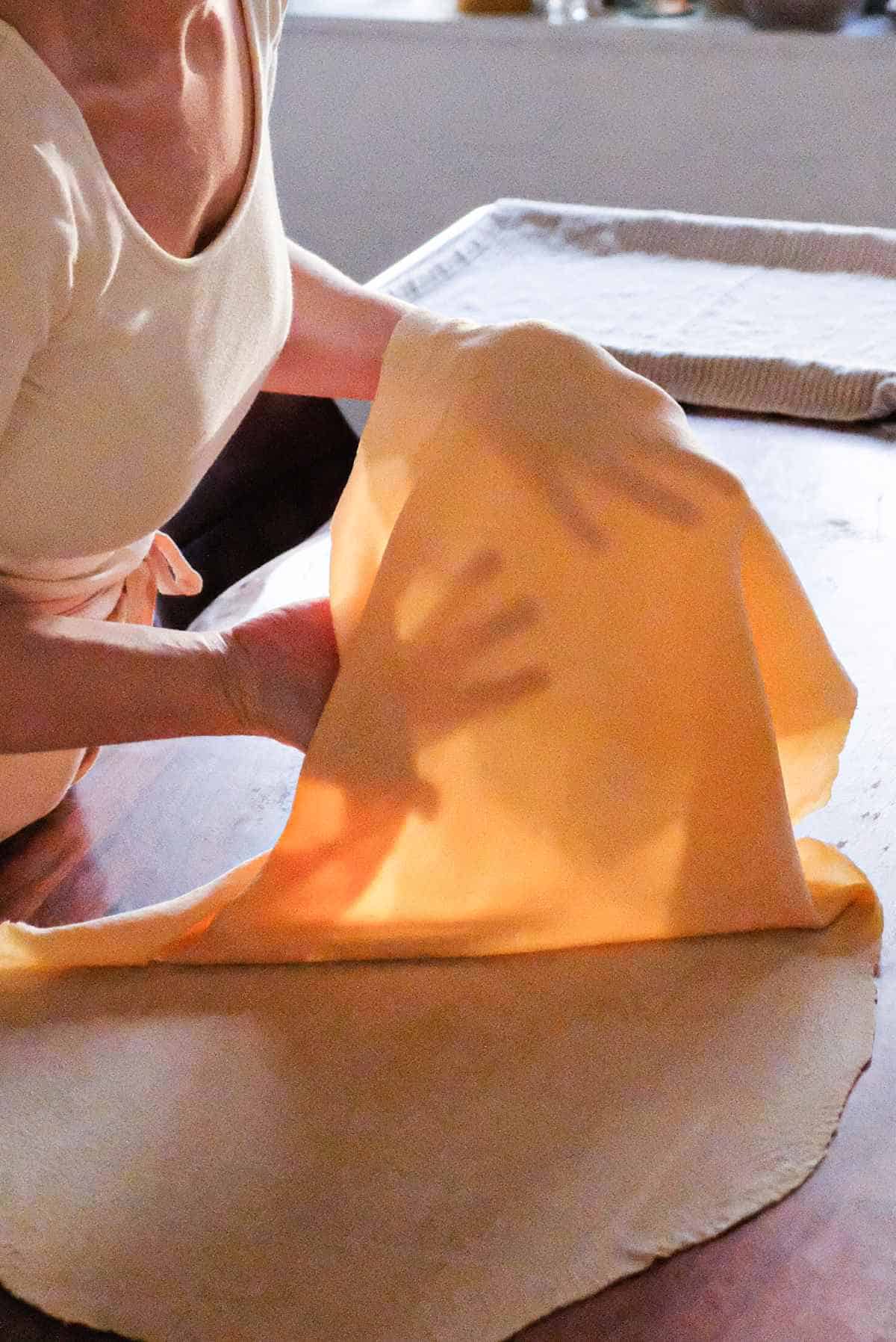 Pasta dough rolled out by hand and lifted up against the light to show that the light can pass through it.