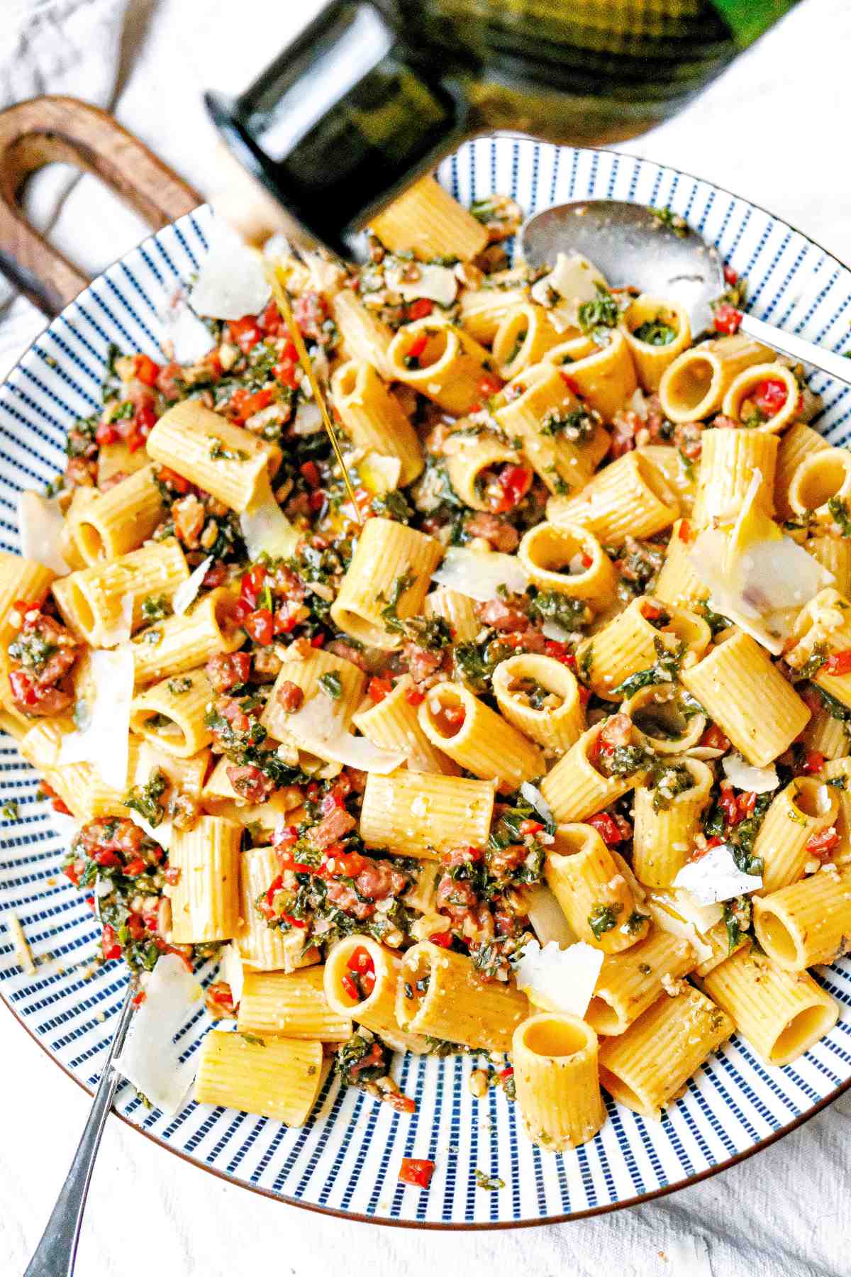 Sausage and kale pasta in a bowl garnished with crumbled walnuts and parmesan petals and being drizzled with olive oil out of a bottle. 