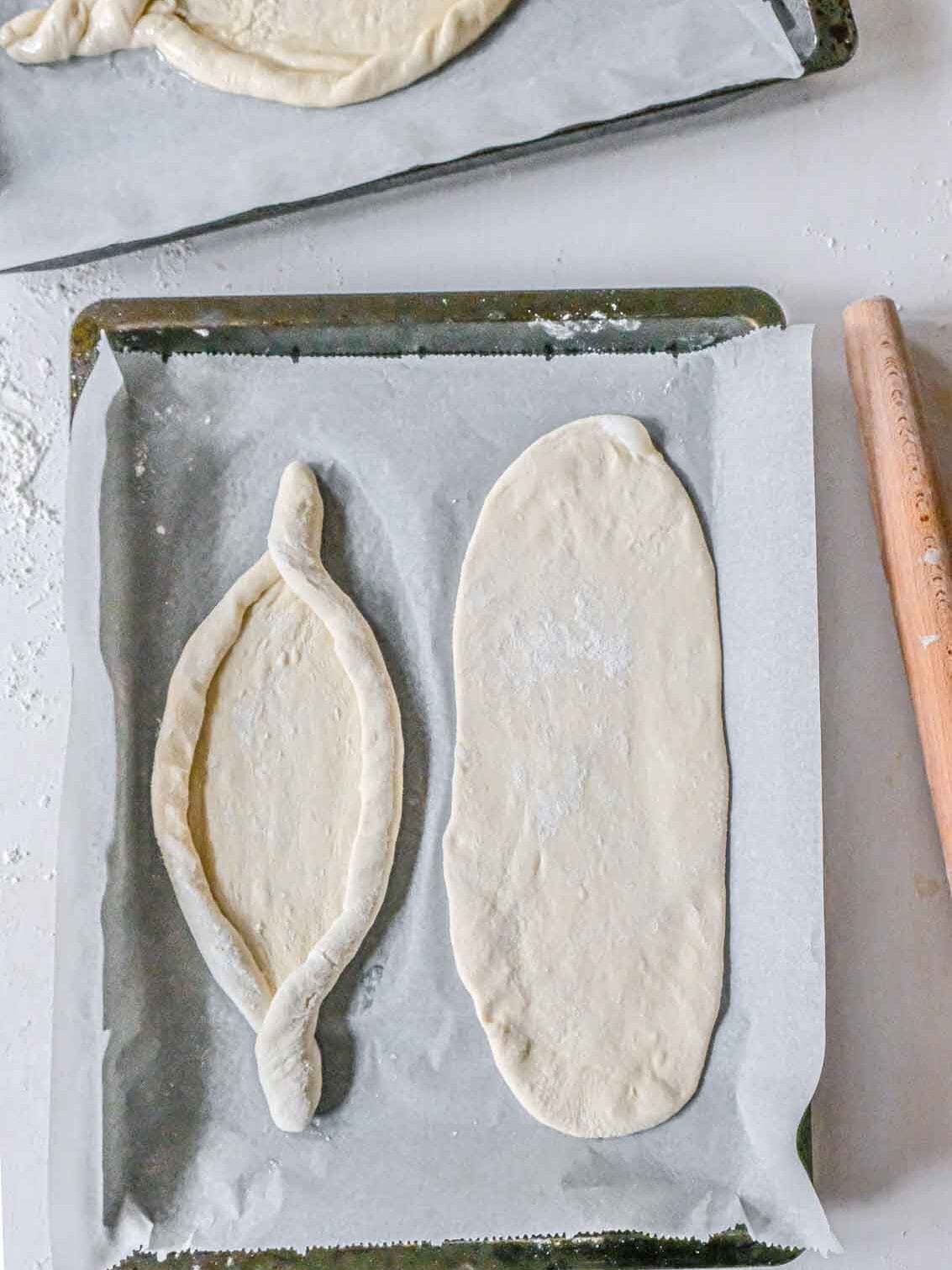 A parchment covered baking tray with one piece of rolled out dough and one piece of rolled out dough shaped into a khachapuri boat ready to be stuffed