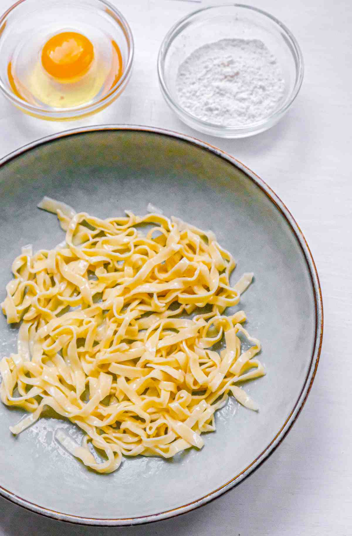 A bowl of fresh tagliatelle pasta with two smaller bowls containing tipo 00 flour and an egg, indicating the ingredients used to make the fresh pasta. 