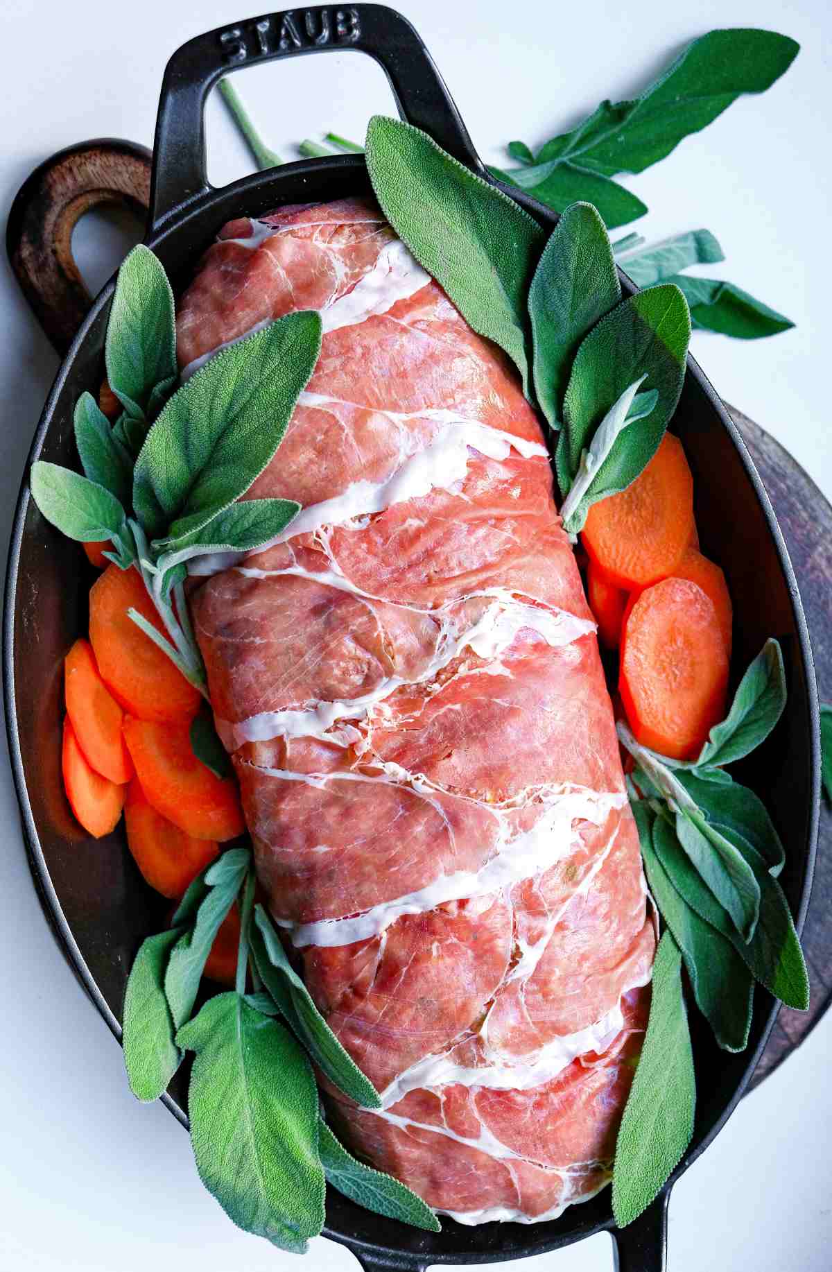 Uncooked meatloaf wrapped in prosciutto in roasting pan with raw sage and carrots.