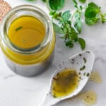 A jar of oregano vinaigrette with a white spoon holding some of the vinaigrette. Some fresh oregano in the background.