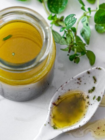 A jar of oregano vinaigrette with a white spoon holding some of the vinaigrette. Some fresh oregano in the background.