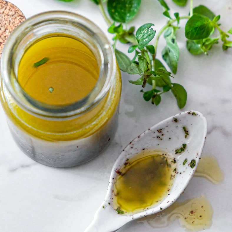 A jar of oregano vinaigrette with a white spoon holding some of the vinaigrette. Some fresh oregano in the background.